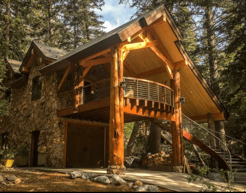 A rustic wooden cabin with a stone facade, featuring a large deck and surrounded by tall trees.